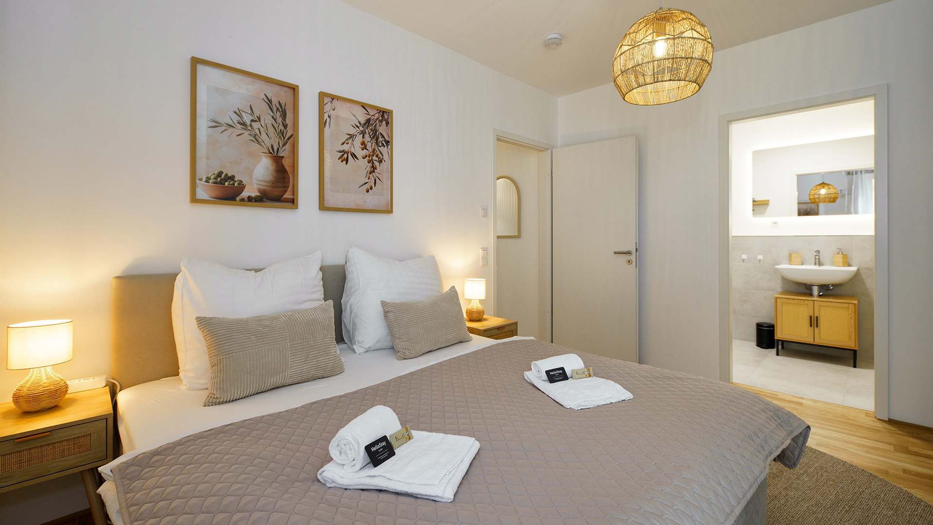 Hotellihuone, jossa on suuri sänky, kaksi yöpöytää, yölamppu, riippuvalaisin, kylpyhuone, jossa on pesuallas ja peili, sekä kaksi kehystettyä taideteosta seinällä.