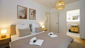 Hotellihuone, jossa on suuri sänky, kaksi yöpöytää, yölamppu, riippuvalaisin, kylpyhuone, jossa on pesuallas ja peili, sekä kaksi kehystettyä taideteosta seinällä.