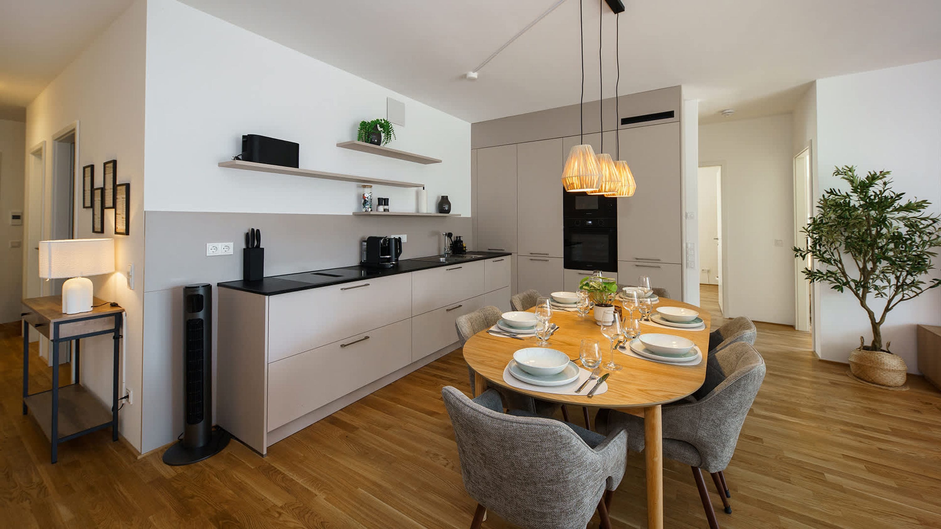 A modern kitchen with a dining area, featuring a wooden table set for six, grey upholstered chairs, a black countertop, and a pendant light.