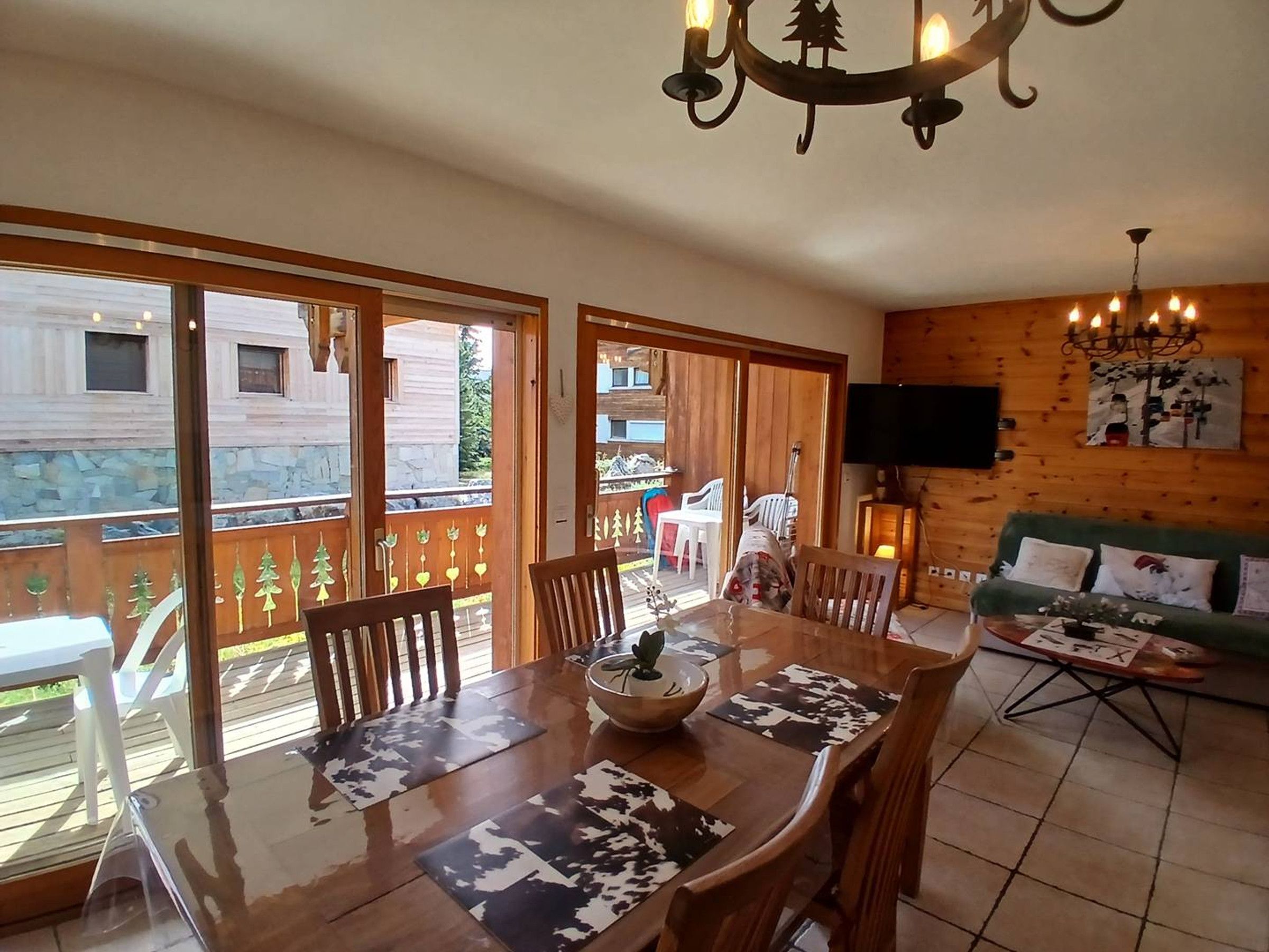 A dining area with a wooden table, chairs, and a chandelier. There is a television mounted on the wall and a sofa with cushions.