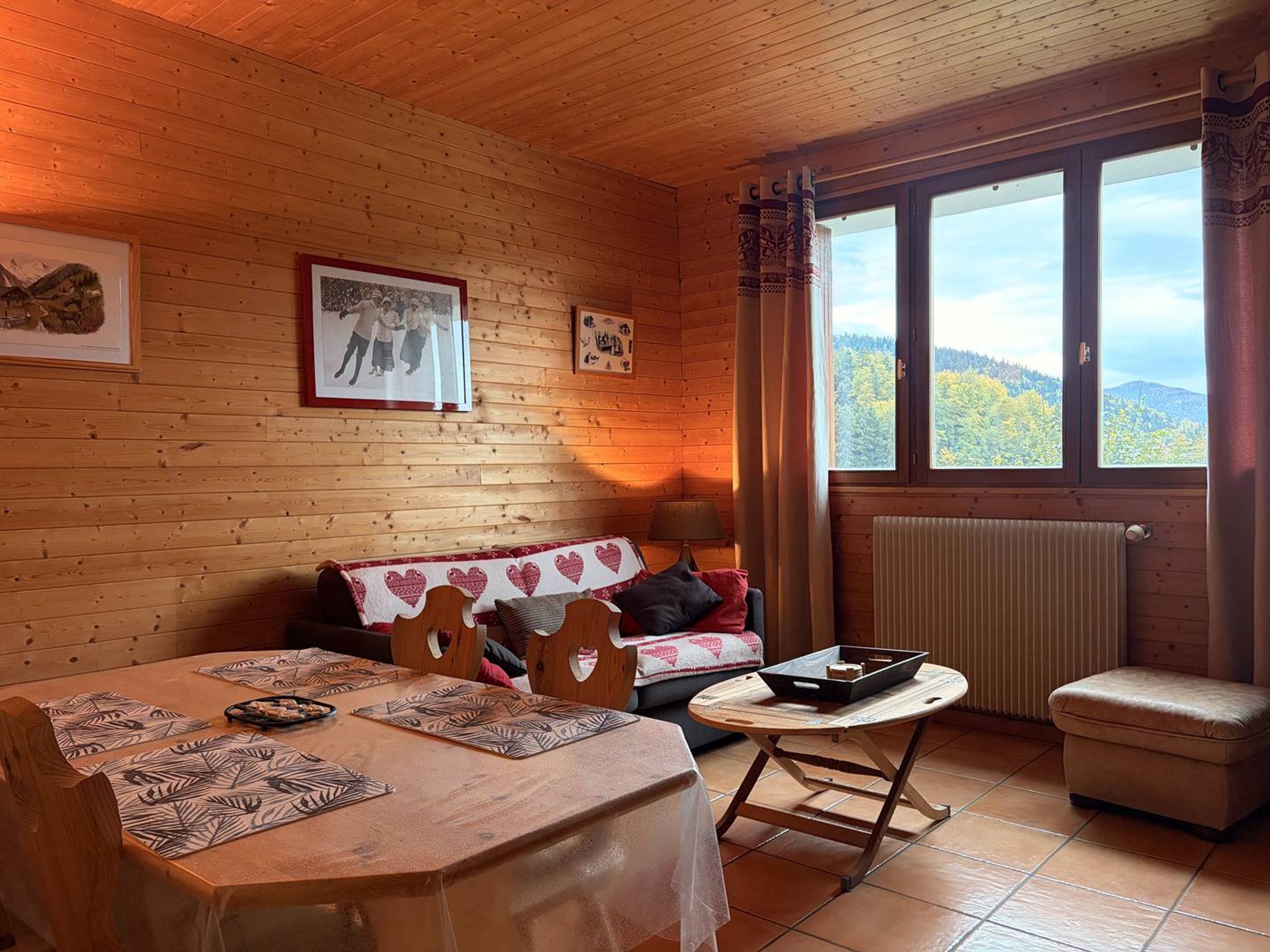 Ein gemütliches Blockhaus-Interieur mit Holzausstattung, einem Esstisch mit Platzsets, einer Couch mit Kissen, einem kleinen Tisch mit Tablett und einem Fenster mit Blick auf den Wald.