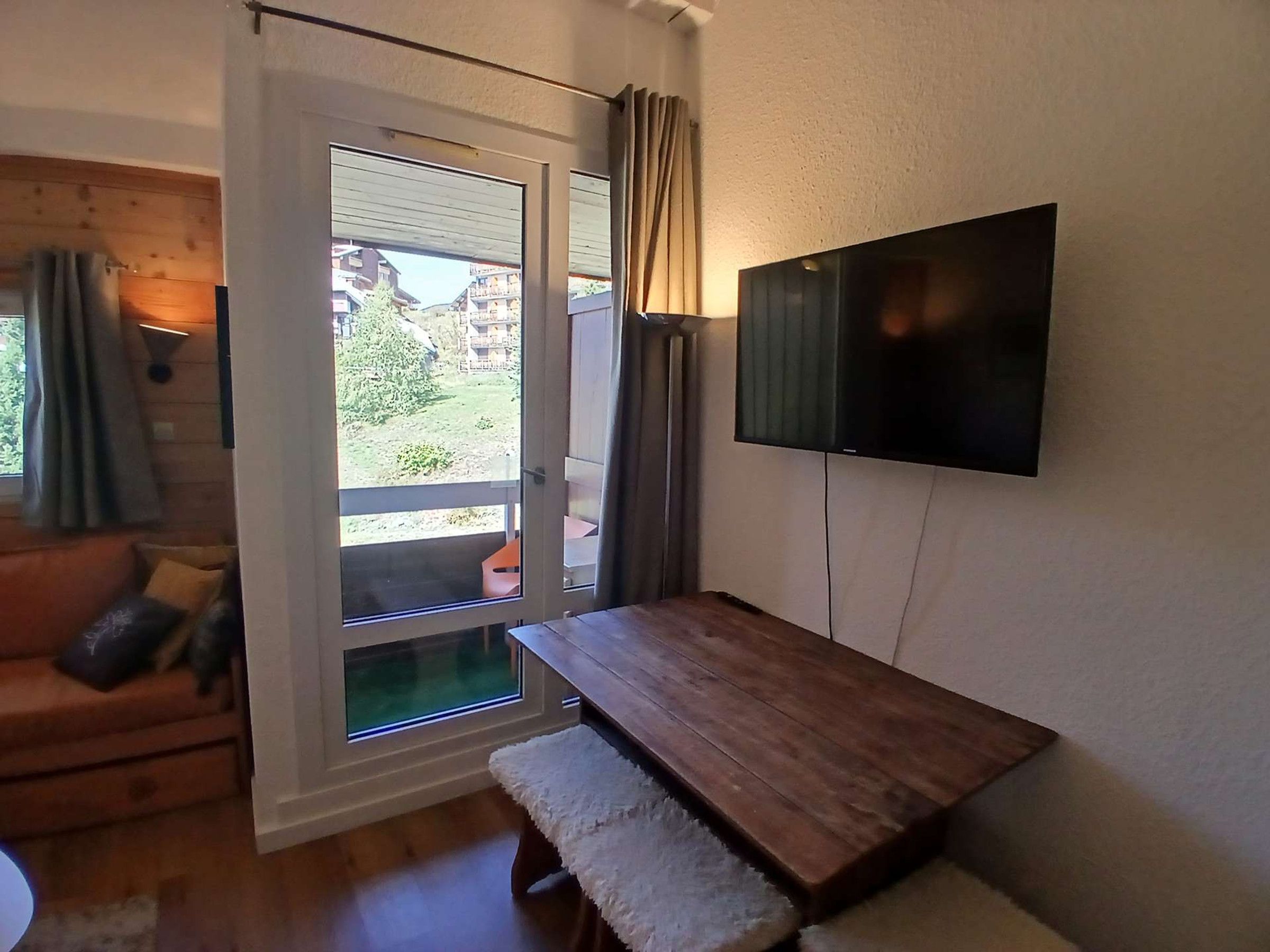 A room with a sliding glass door leading to an outdoor area, a wooden bench, a flat-screen TV mounted on the wall, and a cushioned bench seat.