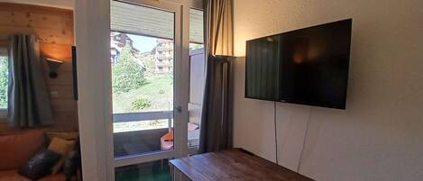 A room with a sliding glass door leading to an outdoor area, a wooden bench, a flat-screen TV mounted on the wall, and a cushioned bench seat.