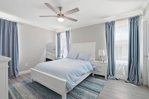 Quarto com cama, mesas de cabeceira, ventoinha no teto e janela com cortinas.
