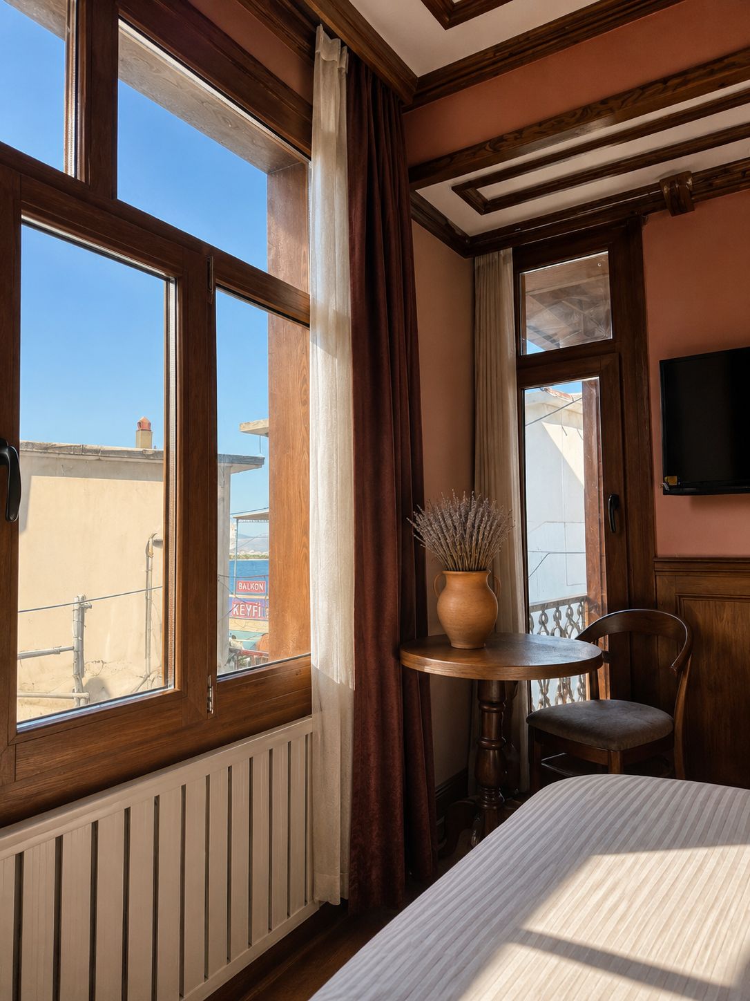 A hotel room with a large window, a bed, a small table with a vase, and a chair.