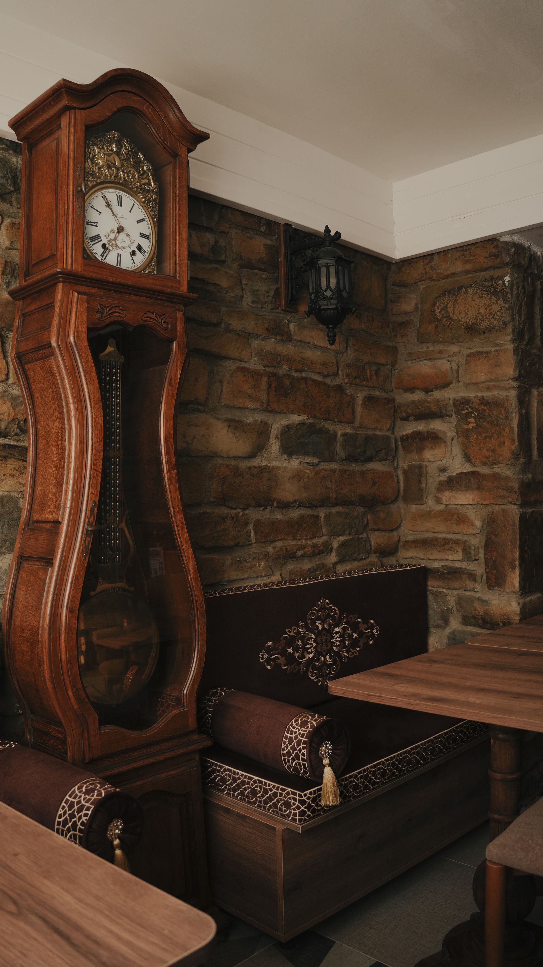 A wooden grandfather clock with Roman numerals, a stone wall, a cushioned bench, and a table.