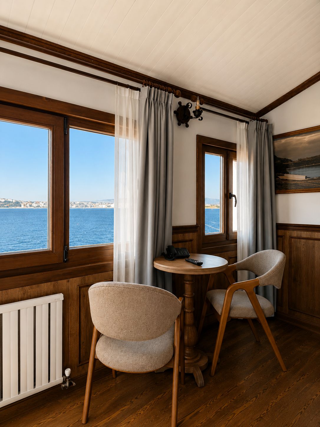 Una habitación con piso de madera, una mesa redonda con dos sillas, un ventanal con cortinas y vista al mar.