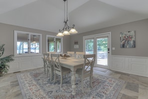 Une salle à manger avec une grande table, des chaises, un lustre et une vue sur l’extérieur, visible à travers les portes vitrées.