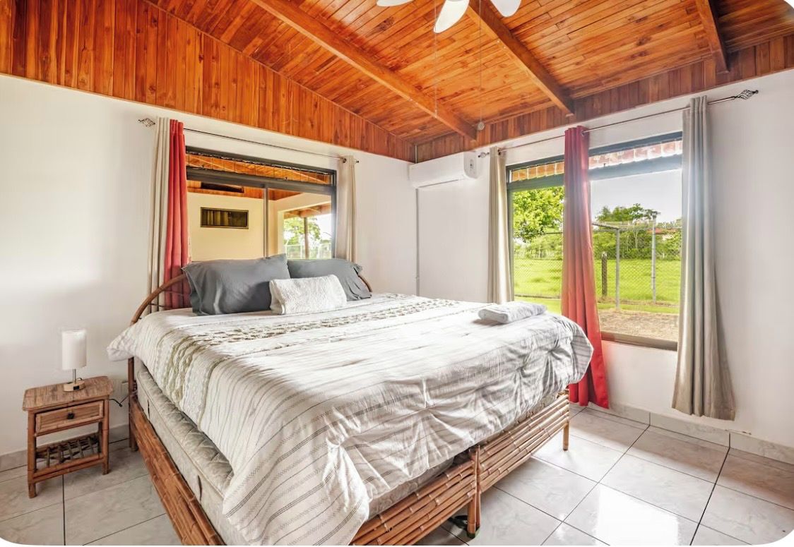 Une chambre avec un plafond en bois, un lit recouvert d’une couvre-lit à rayures blanches et grises, une table de chevet avec une lampe et une fenêtre avec des rideaux.
