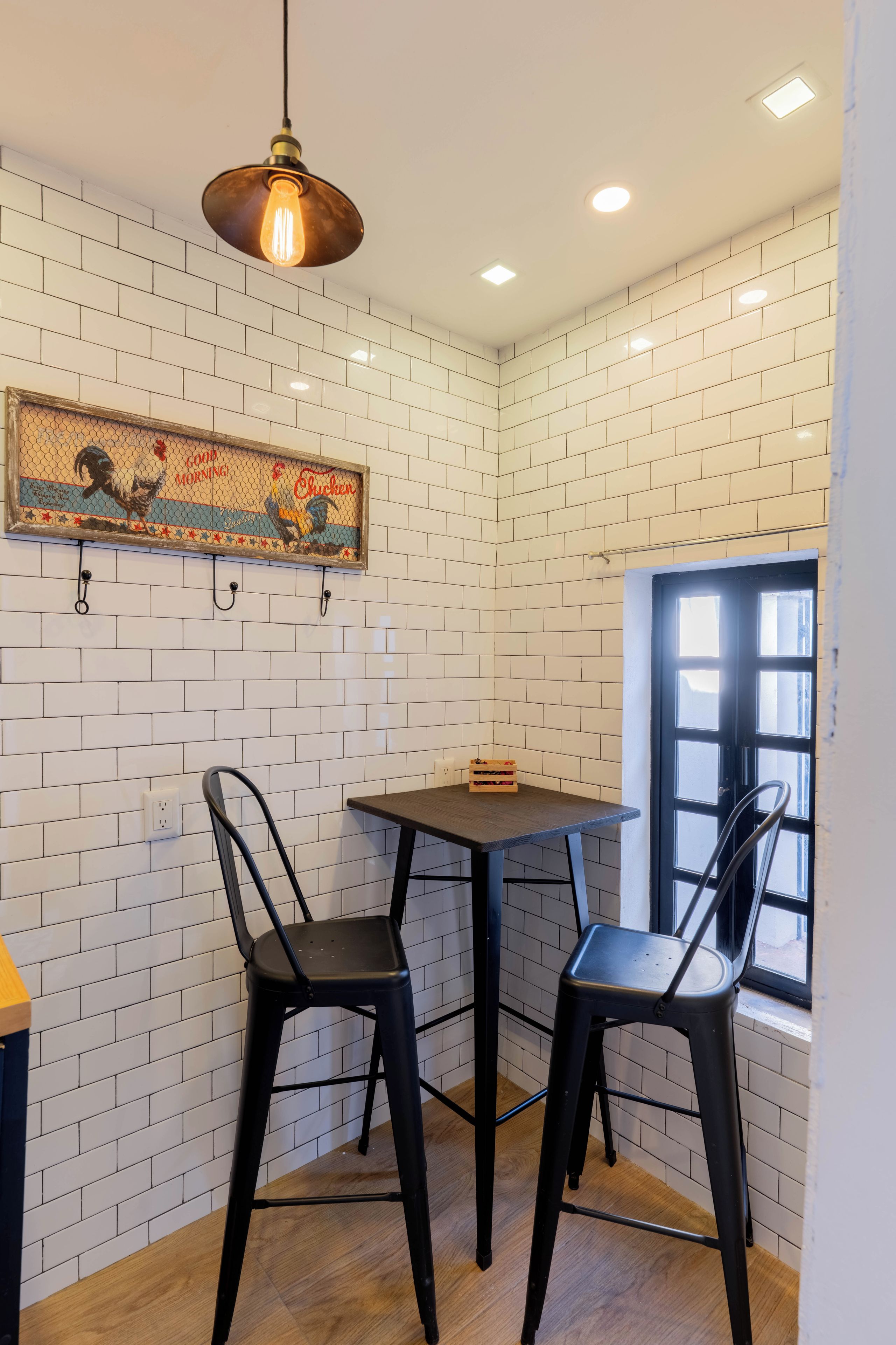 A small dining area with two black chairs, a wooden table, a hanging light fixture, and a wall-mounted picture.