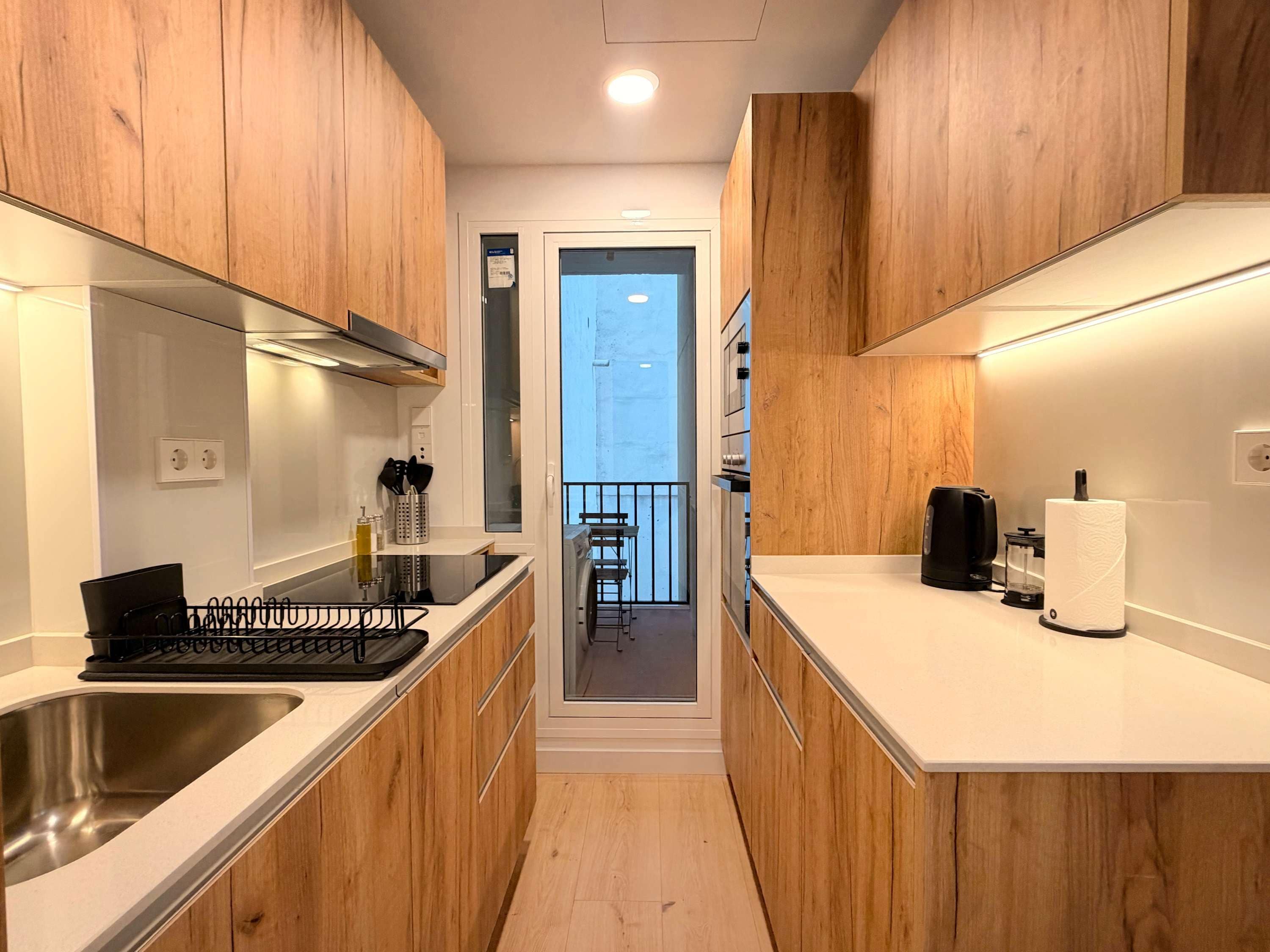 A narrow kitchen with wooden cabinets, a stainless steel sink, and a black electric kettle.