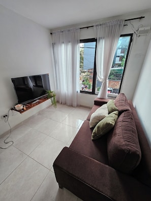 A living room with a flat-screen TV mounted on the wall, a brown sofa with cushions, a small wooden shelf with a plant, and a window with white curtains.
