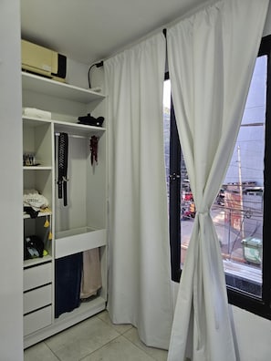 A room with a white wardrobe, a window with white curtains tied back, and a view of the street outside.