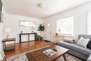 Una sala moderna con un sofá, una mesa de centro y una mesita auxiliar. Hay una puerta con un espejo encima, y una ventana con persianas.