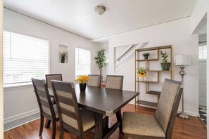 Comedor con una mesa de madera oscura, seis sillas y un jarrón con flores amarillas. Hay dos ventanas con persianas, una planta en el alféizar y una lámpara de pie.