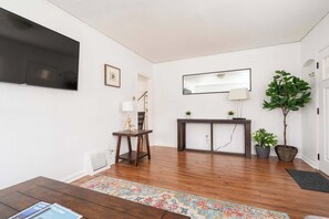 Una sala de estar moderna con una televisión de pantalla plana, una mesa de centro de madera, una consola con plantas, un espejo grande y un árbol en maceta.