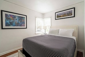 Un dormitorio con una cama, dos cuadros enmarcados, una ventana con persianas y una lámpara de noche.
