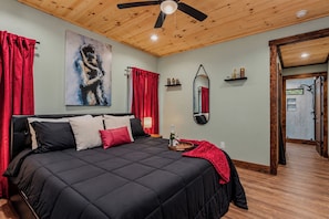 Une chambre à coucher avec un lit, un ventilateur de plafond, un tableau, un miroir et un rideau rouge.
