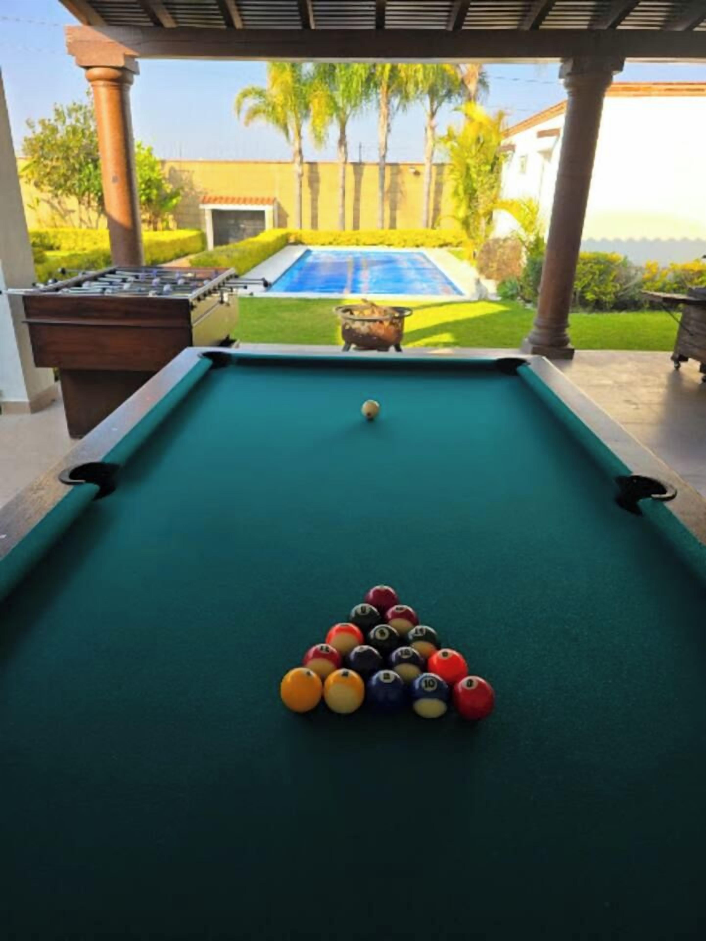 Une table de billard avec les billes disposées en forme de triangle, une table de billard située dans un espace abrité, avec vue sur une piscine et des palmiers.