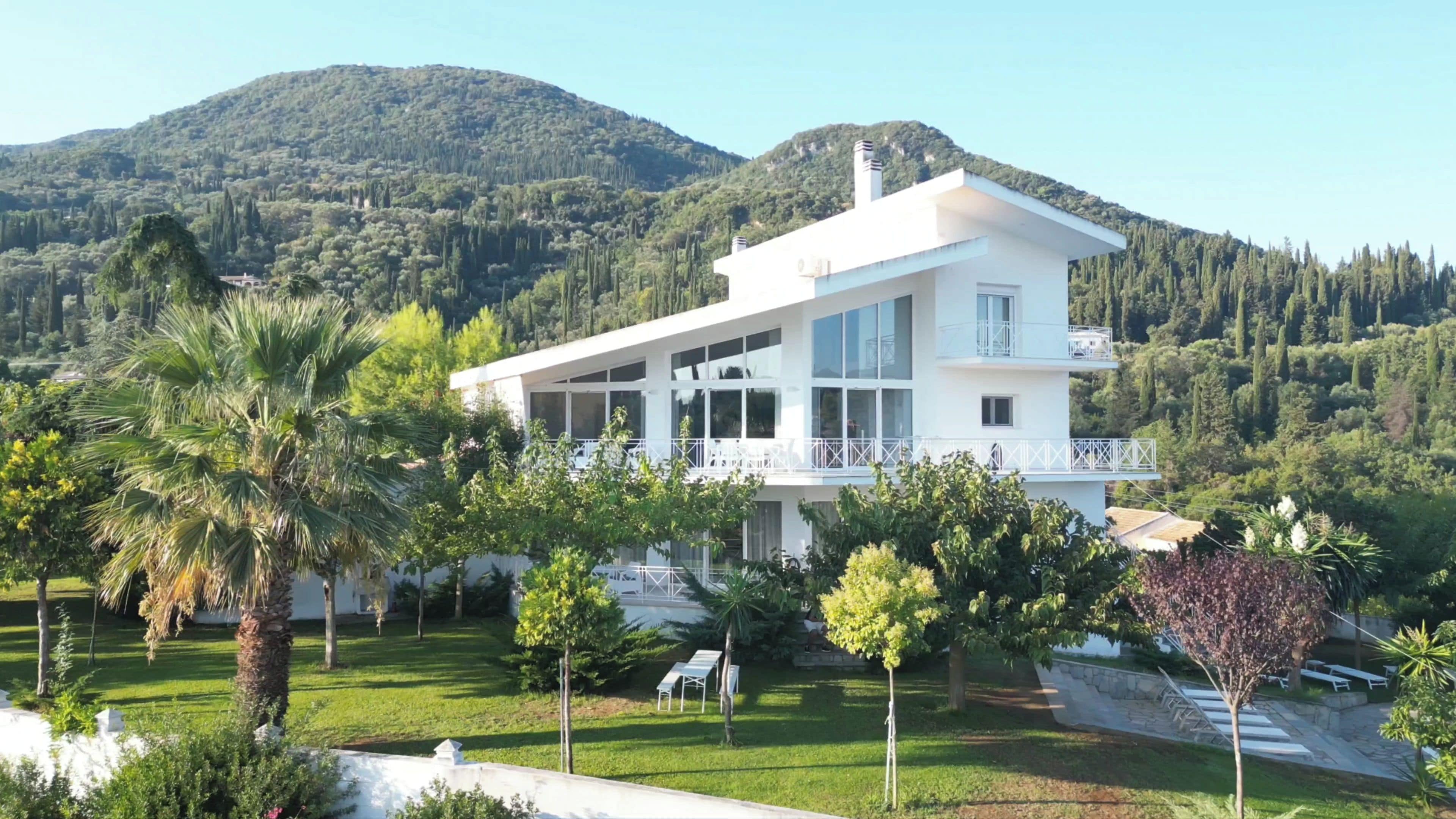 A modern two-story house with a balcony, surrounded by lush greenery and a mountain in the background.