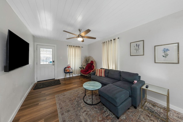 A living room with a sofa, a small round table, a television, a ceiling fan, and a door with a window. - Modern & Bright 2BR Home Near East Lake Park (Birmingham)
