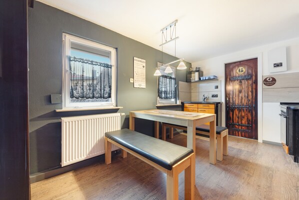 Een kleine keuken met een eethoek, een radiator, een houten deur en een raam met gordijnen.