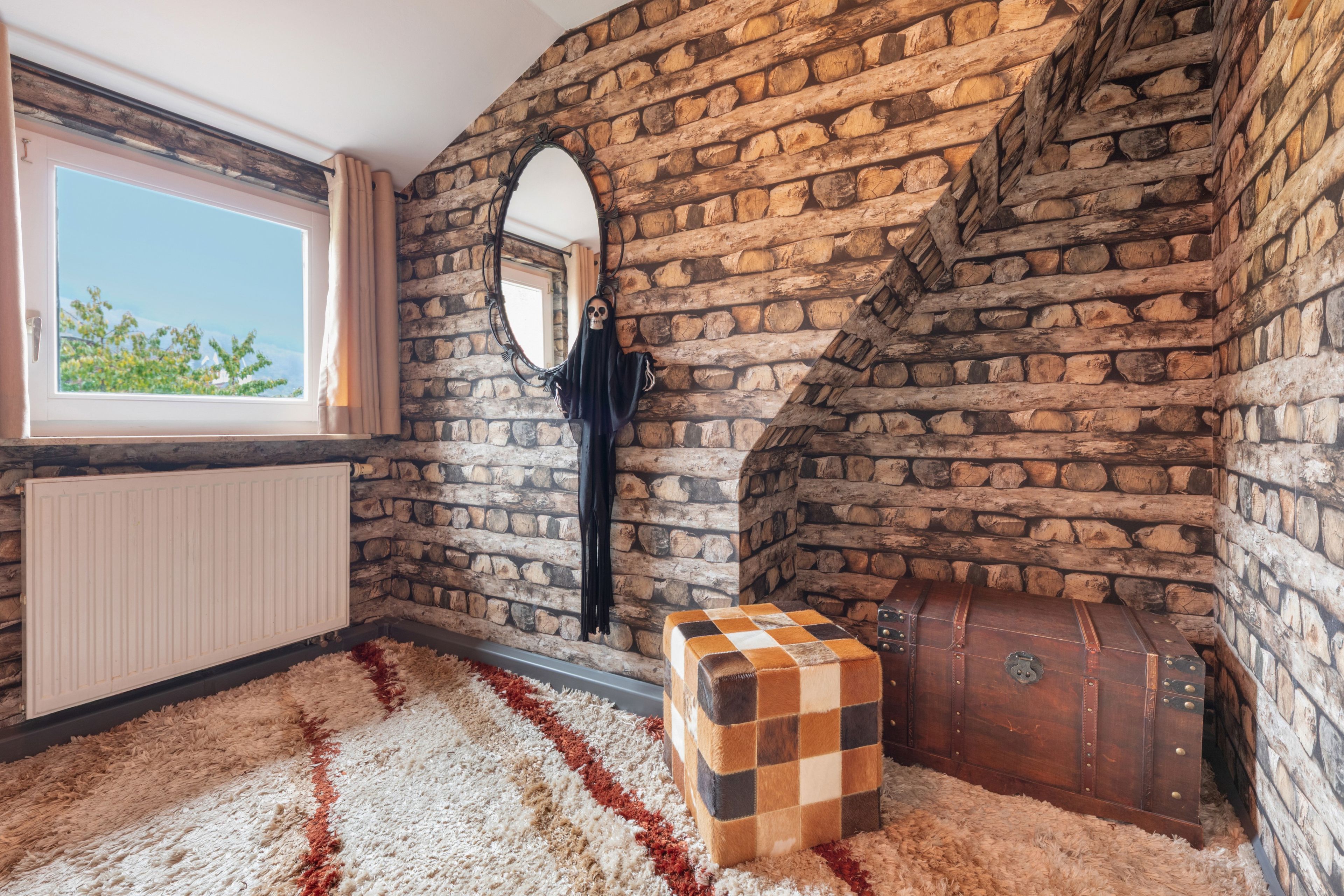 A room with a stone wall, a window with curtains, a radiator, a checkered ottoman, and a trunk.