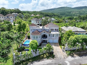 Uma casa grande, com vários andares, e uma piscina, rodeada por vegetação exuberante e colinas ao fundo.