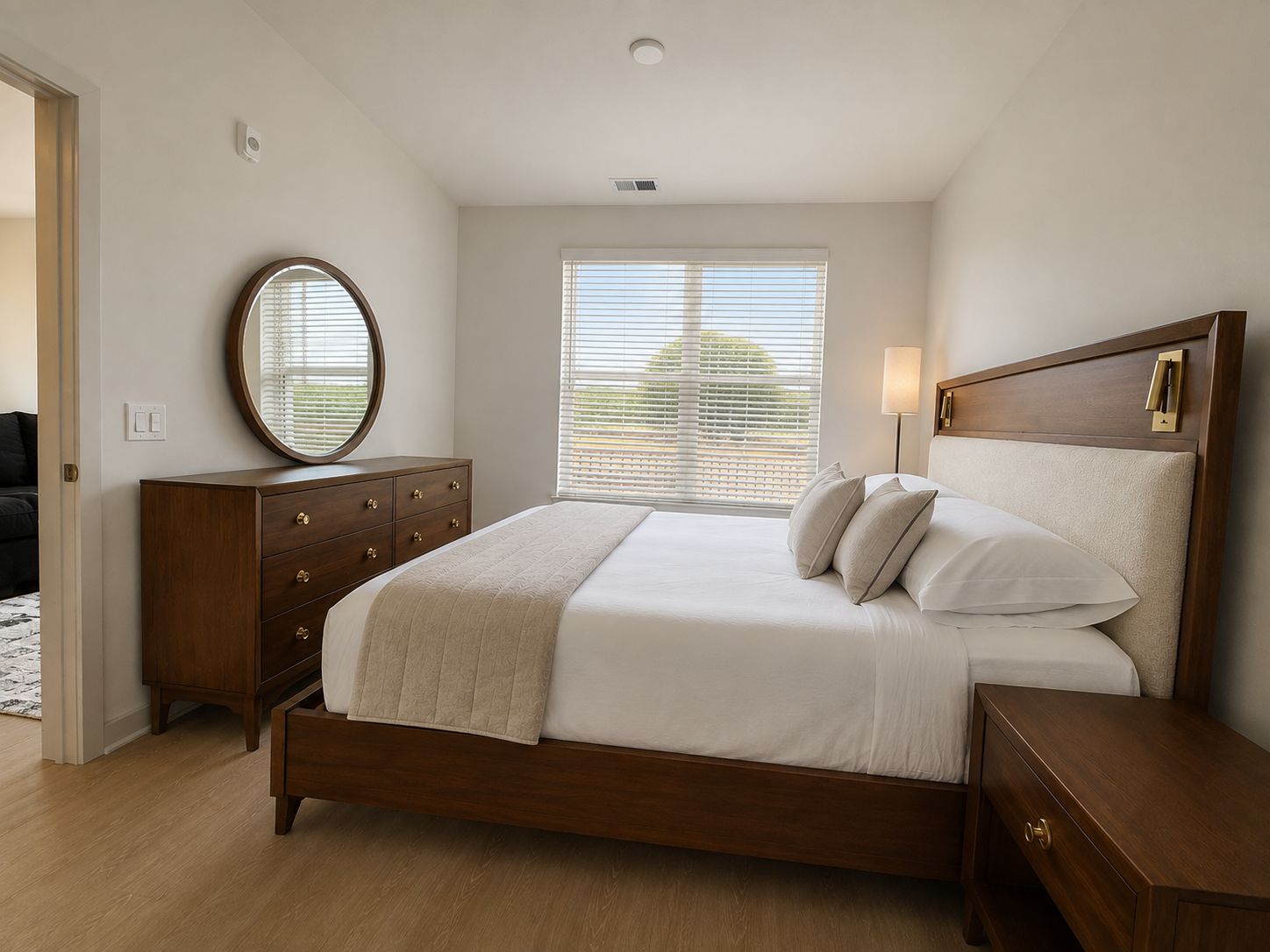 A bedroom with a wooden bed, a dresser, a bedside table, a lamp, and a window with blinds.