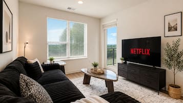 A living room with a black sofa, a coffee table, a TV displaying 'NETFLIX', and a balcony with a view.