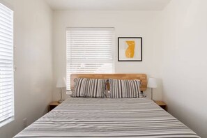 Ein Schlafzimmer mit einem Bett, einem Holz-Kopfteil, zwei Nachttischlampen, einem gerahmten Bild an der Wand und einem Fenster mit Jalousien.