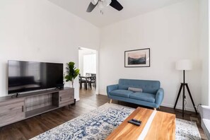 A modern living room with a flat-screen TV, a blue sofa, a wooden coffee table, and a floor lamp. - Division St 3BR • Tilikum Bridge Views • Sleeps 6 (Portland)
