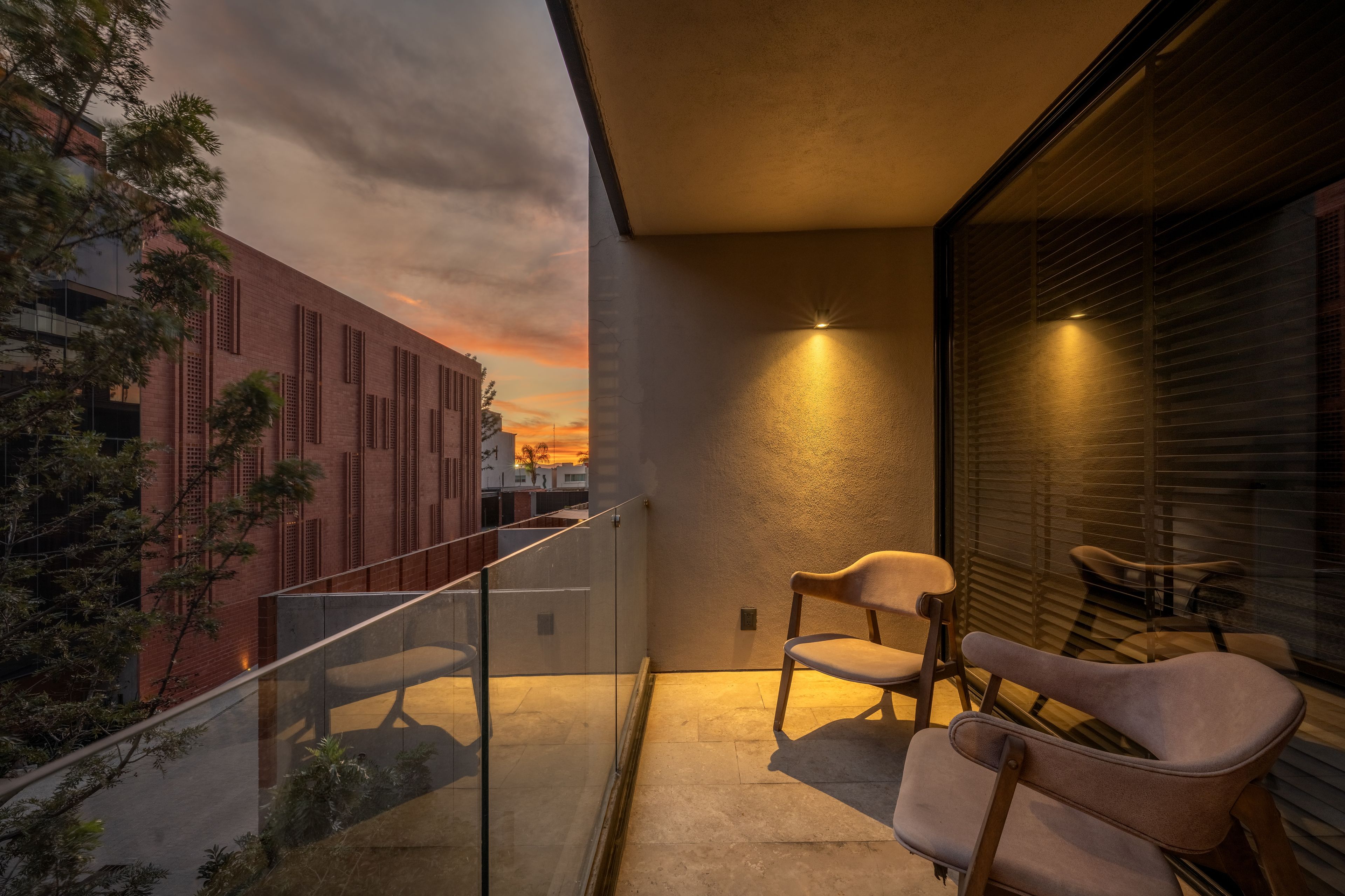 A balcony with a glass railing, two chairs, a small table, and a view of a sunset over a cityscape.