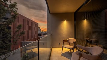 A balcony with a glass railing, two chairs, a small table, and a view of a sunset over a cityscape.
