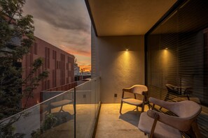 Ein Balkon mit Glastrennwand, zwei Stühle, ein kleiner Tisch und Blick auf einen Sonnenuntergang über einer Stadtlandschaft.