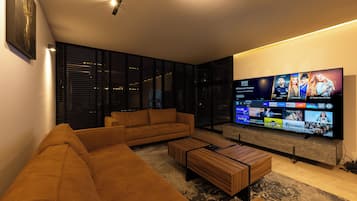 A modern living room with a large flat-screen TV, a brown sofa, a wooden coffee table, and a glass partition with blinds.