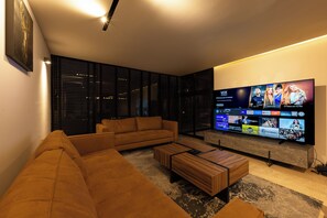 Ein modernes Wohnzimmer mit einem großen Flachbildfernseher, einem braunen Sofa, einem Holztisch und einer Glastrennwand mit Jalousien.