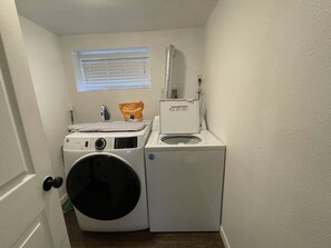 Ein kleiner Hauswirtschaftsraum mit einer Waschmaschine und einem Trockner mit Frontlader, einem Fenster mit Jalousien und einem Abluftventilator am Trockner.