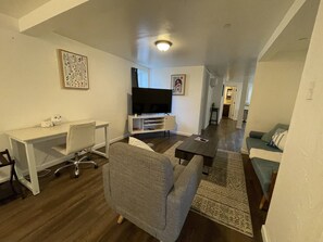 Ein modernes Wohnzimmer mit Flachbildfernseher, grauem Sofa, weißem Schreibtisch und einem gemusterten Teppich.