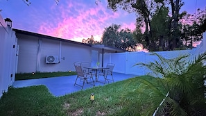 A patio with a table and chairs, a white fence, and a lawn.