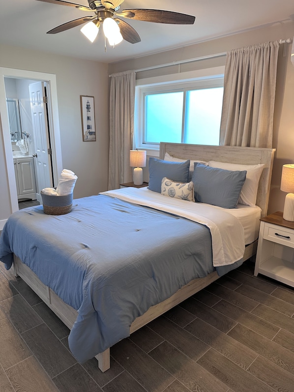 A bedroom with a bed, a nightstand, a ceiling fan, a window with curtains, and a bathroom visible in the background.