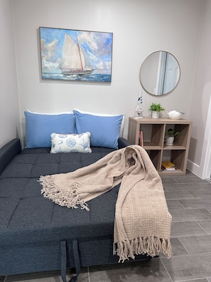 A modern bedroom with a dark grey sofa, blue pillows, a beige throw blanket, a painting of a sailboat, a round mirror, and a small plant on a wooden shelf.