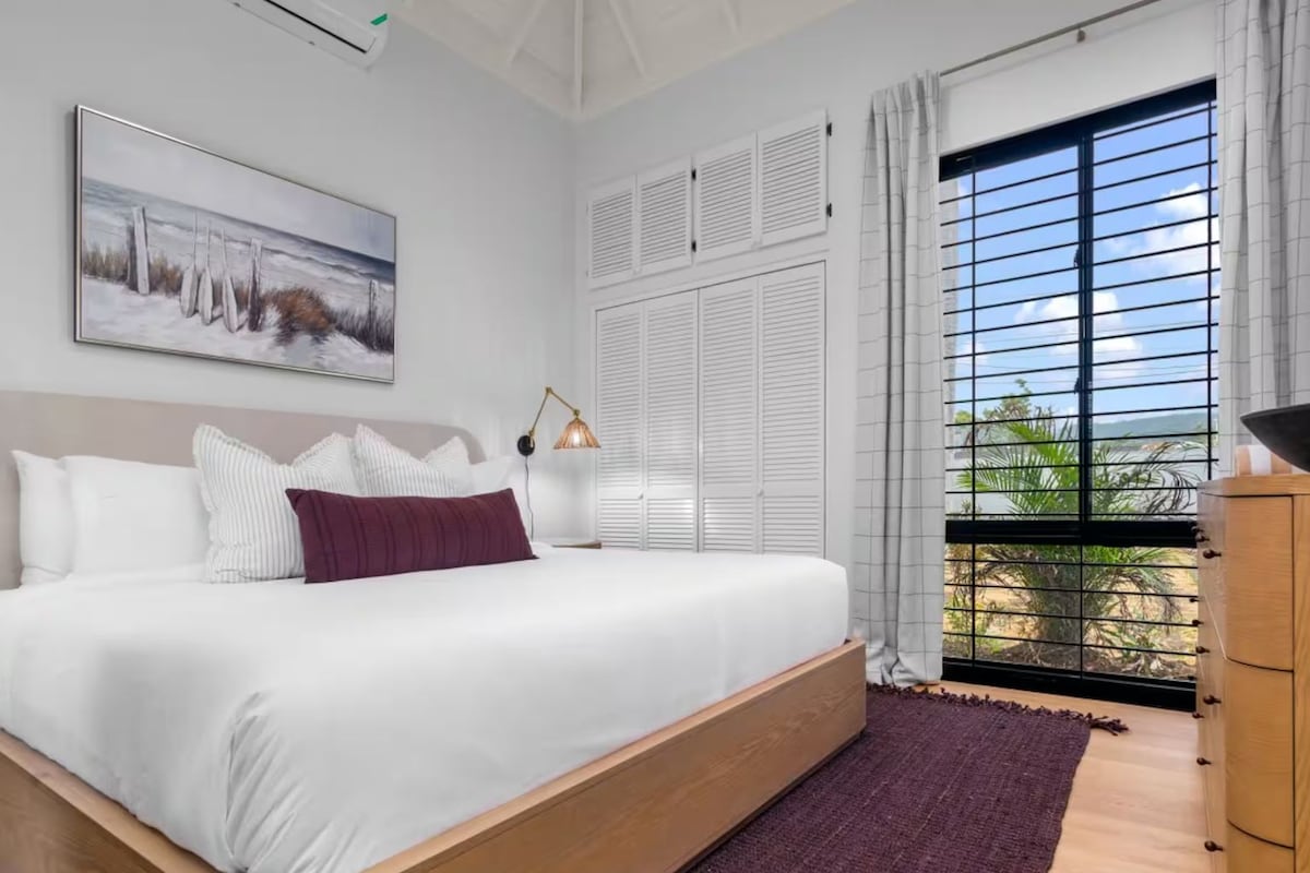 A bedroom with a large bed, a painting on the wall, a window with shutters, and a view of the outdoors.
