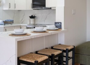 A kitchen with a white countertop, three bar stools with woven seats, and a microwave above.