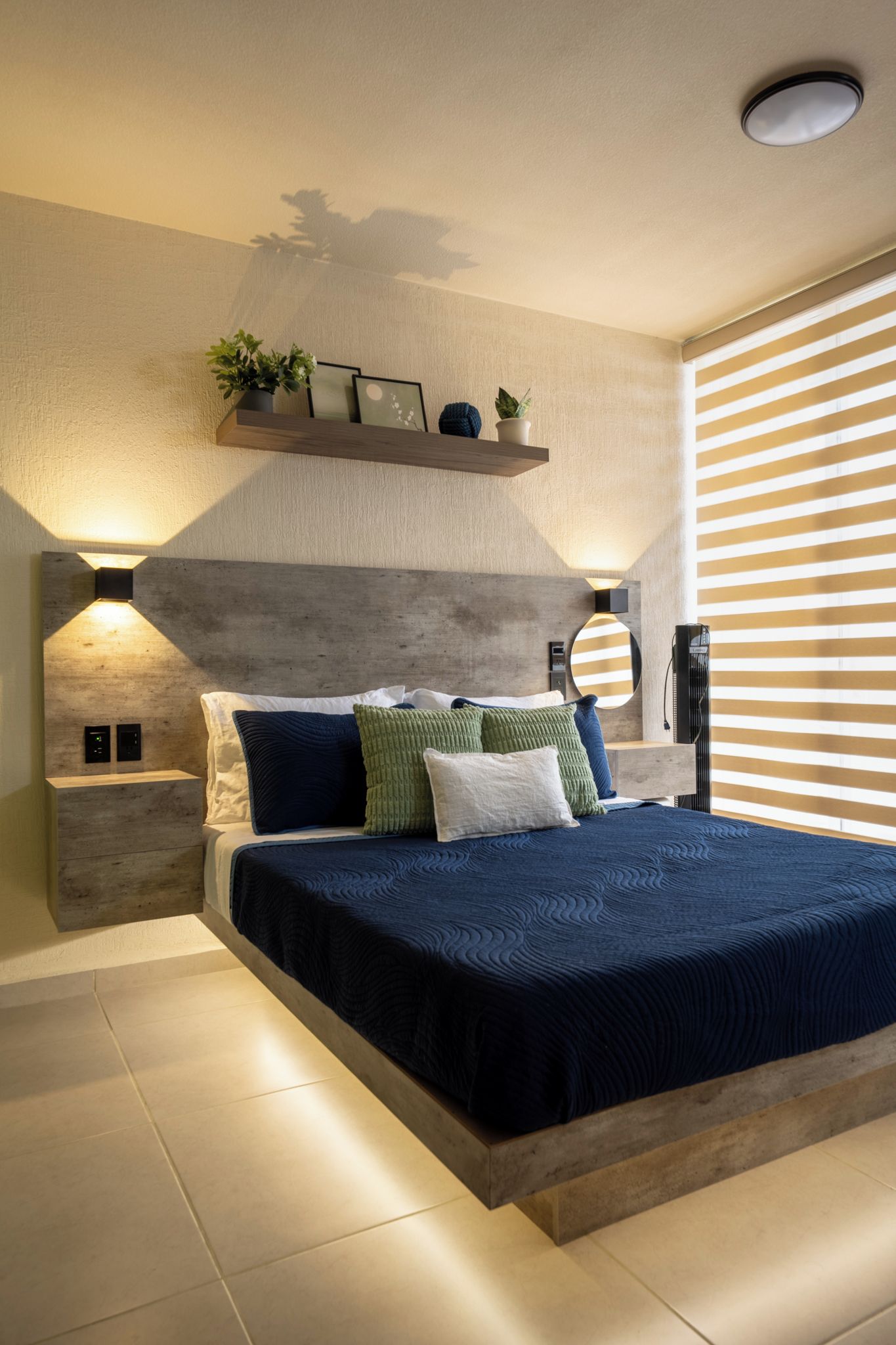 A modern bedroom with a large bed, bedside tables, a wall-mounted shelf with plants, a round mirror, and a ceiling light fixture.