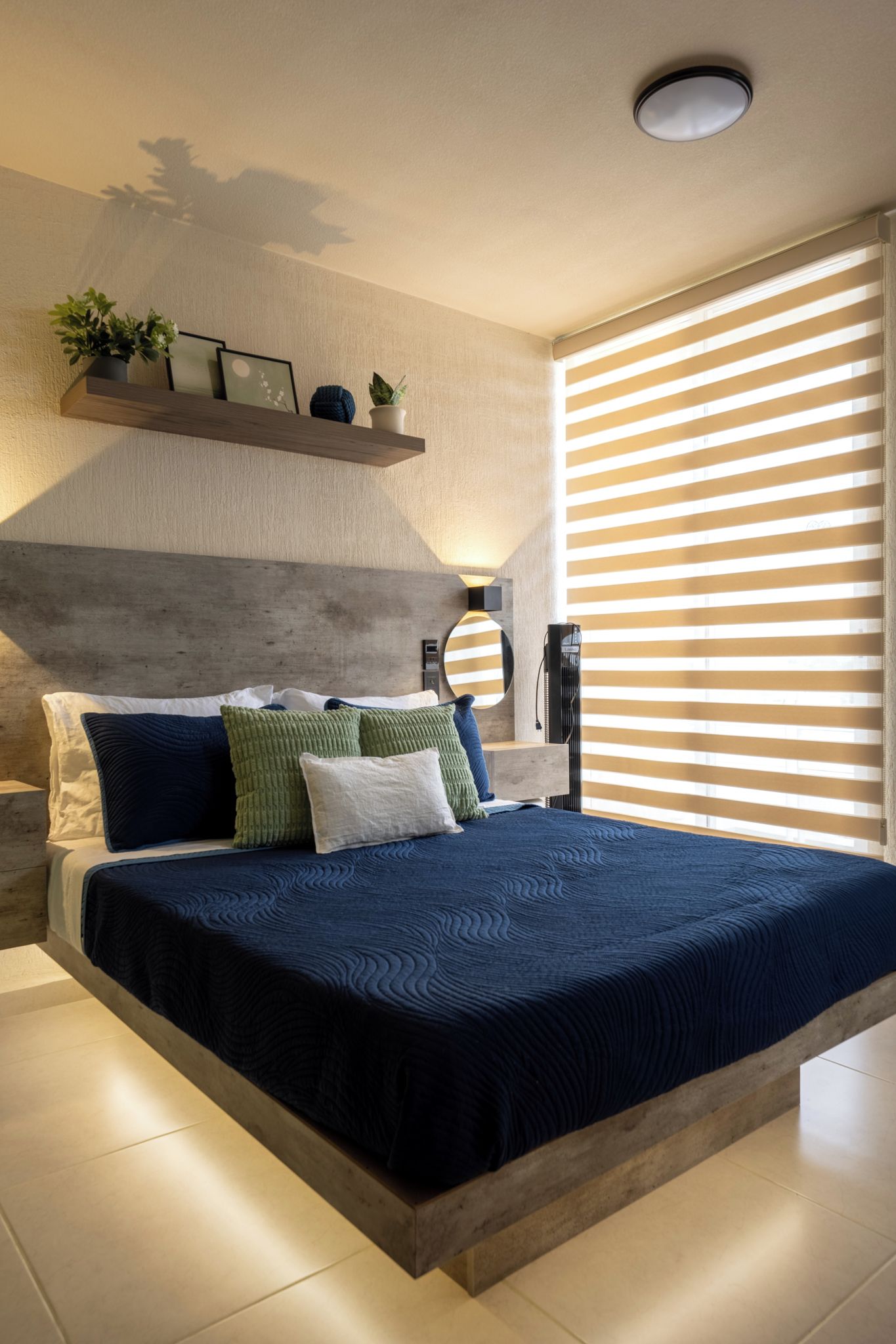 Une chambre moderne avec un grand lit, une tête de lit en bois, une table de chevet, une étagère murale agrémentée de plantes et une fenêtre avec des stores horizontaux.