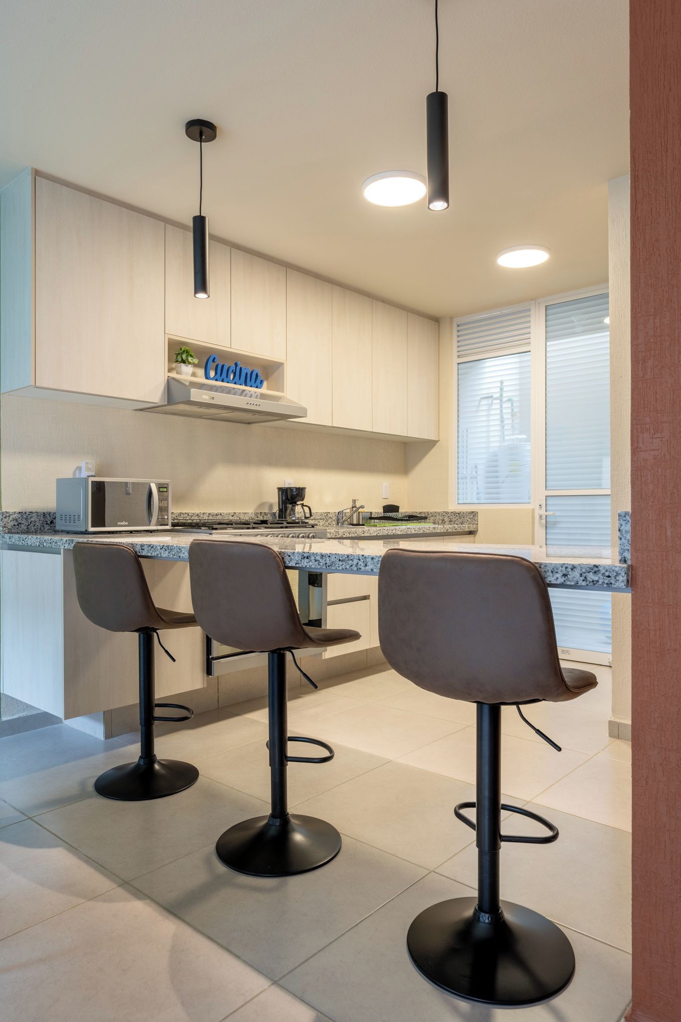 A modern kitchen with a microwave, a coffee maker, and a sink. There are three bar stools and a ceiling light.