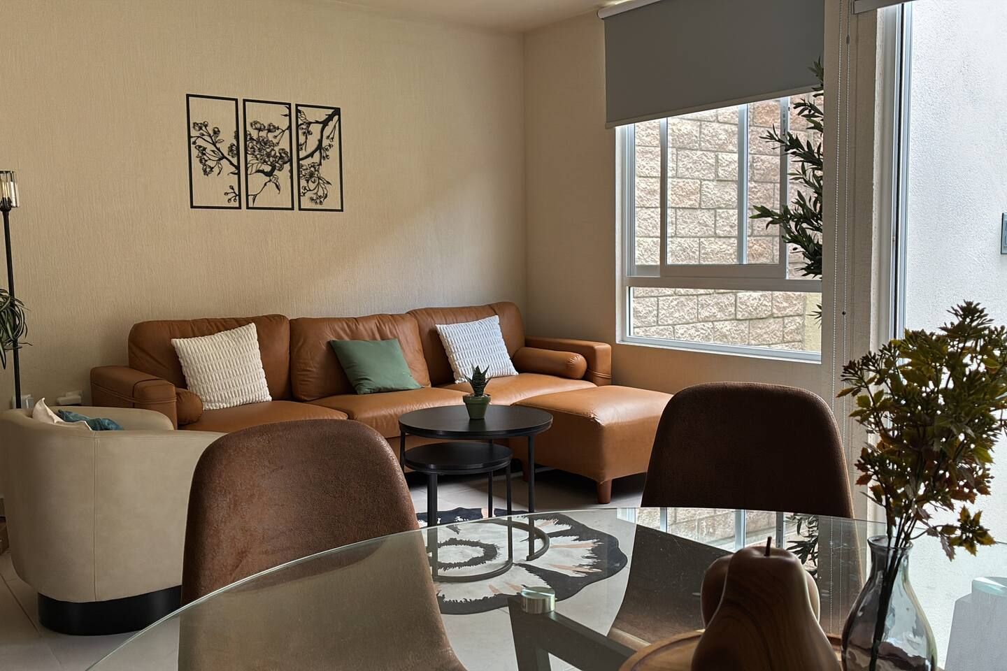 Un salon avec un canapé en cuir marron, une table basse en verre et un fauteuil. Trois tableaux encadrés ornent le mur, et une fenêtre offre une vue sur les arbres.