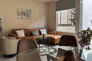 Un salon avec un canapé en cuir marron, une table basse en verre et un fauteuil. Trois tableaux encadrés ornent le mur, et une fenêtre offre une vue sur les arbres.
