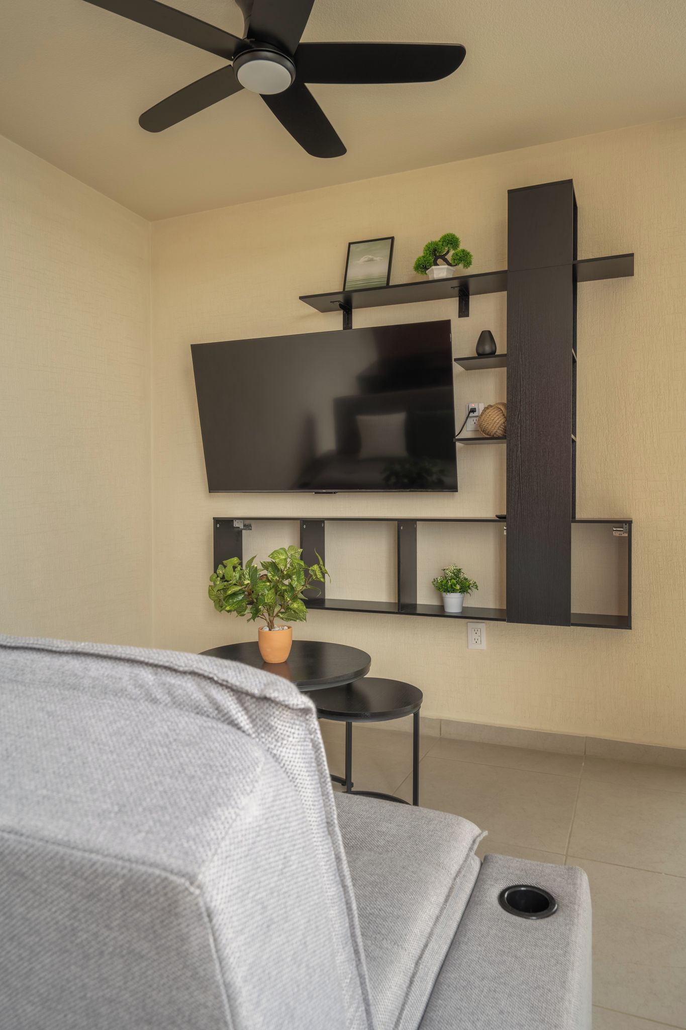 Ein Wohnzimmer mit Flachbildfernseher an der Wand, Deckenventilator und grauem Sofa.